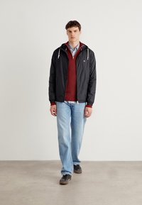 Sort zip-up jakke med hætte over en bordeaux strikket sweater, parret med lyseblå jeans og mørke sneakers. Enkel, afslappet design.