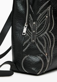 Zaino in pelle nera con superficie testurizzata, zip argentata, dettagli ricamati bianchi e accenti metallici per decorazione.