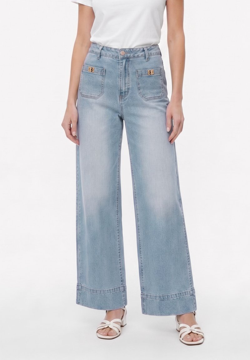 Femme portant un jean bleu clair taille haute à jambes larges avec des poches plaquées à l'avant et un haut blanc à manches courtes, associé à des sandales blanches ouvertes aux orteils.