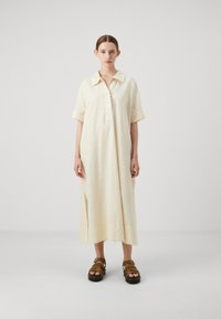 Vestito in lino giallo chiaro con collo a camicia, maniche corte e spacchi sui lati. Abbinato a sandali marroni con fibbie.