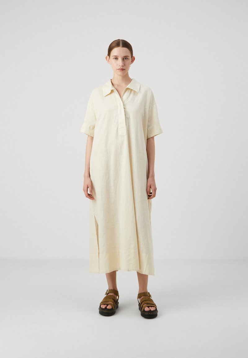 Vestito in lino giallo chiaro con collo a camicia, maniche corte e spacchi sui lati. Abbinato a sandali marroni con fibbie.