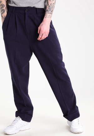 Personne portant un pantalon plissé bleu marine et des baskets blanches, debout avec une main détendue le long du corps et des tatouages visibles sur les bras.