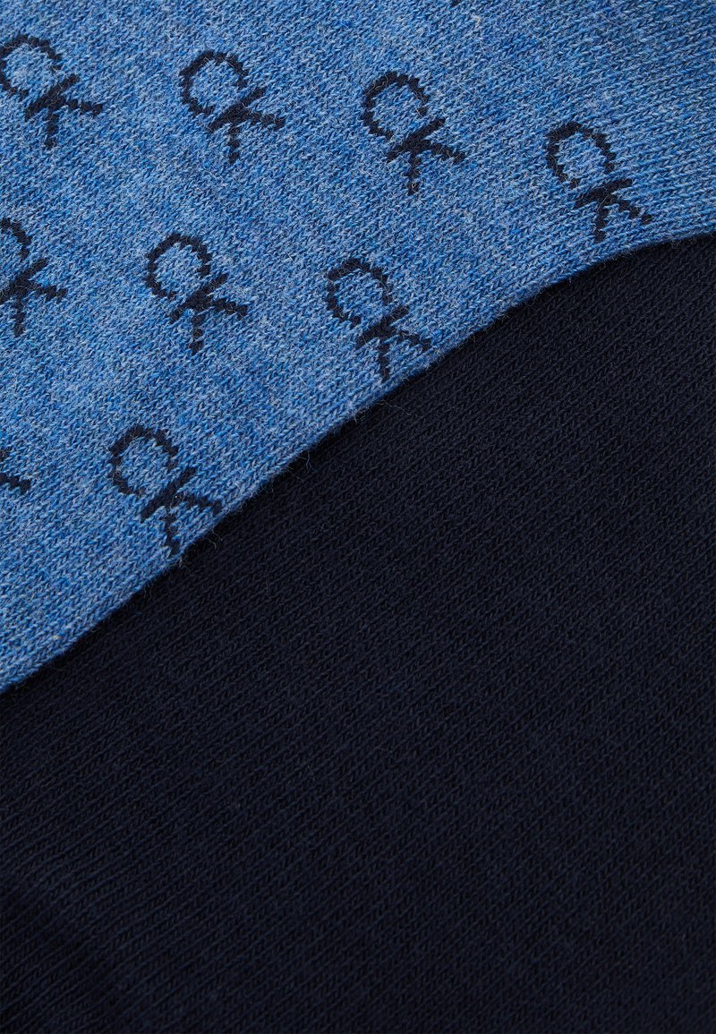 Calvin Klein Socken mit einem blauen Strickbereich, der wiederholt schwarze "CK"-Logos zeigt, und einem kontrastierenden einfarbigen schwarzen Strickbereich.