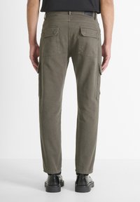 Uomo che indossa pantaloni cargo dritti color verde oliva e stivali di pelle neri, in piedi di spalle contro uno sfondo bianco uniforme.