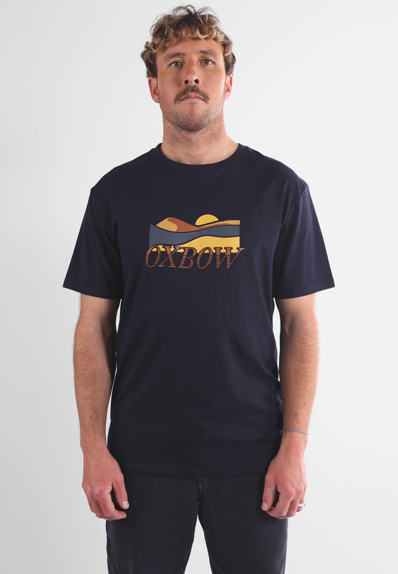 Homme aux cheveux frisés et à la moustache se tient face à l'avant, portant un t-shirt bleu marine avec un paysage de coucher de soleil et le texte "OXBOW".