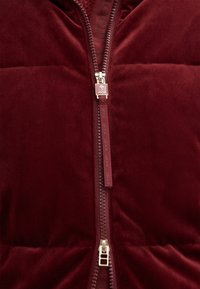 Il gilet imbottito bordeaux presenta una texture vellutata liscia, una cerniera a contrasto e un tirazip con logo. 