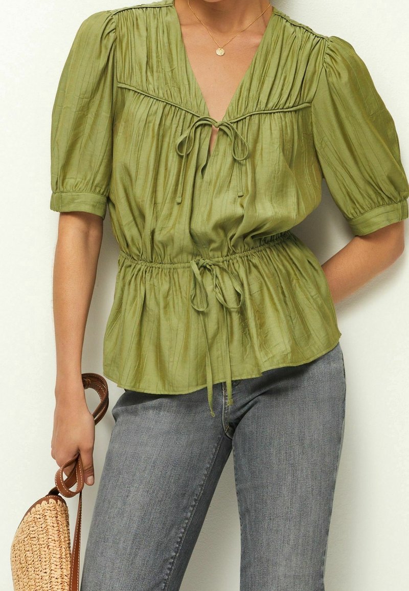 Blouse texturée vert olive avec manches bouffantes, encolure en V et taille cintrée avec un détail noué. Portée avec un jean gris et un sac en paille.