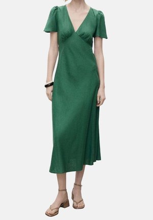 Femme portant une robe midi verte à encolure en V, manches courtes bouffantes et sandales marron clair à talon bloc et brides, debout sur un fond uni.
