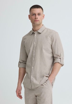 Homme aux cheveux courts portant une chemise beige à boutons avec manches retroussées et pantalon assorti, debout avec une main dans la poche sur un fond uni.