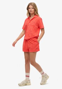 Chemise courte à manches courtes couleur corail et shorts assortis avec des motifs de feuilles texturés. Les shorts ont une taille élastique et des poches latérales. Porté avec des baskets beiges.