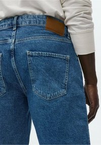 Jeans en denim bleu avec une finition texturée, featuring une patch en cuir marron à la taille et deux poches arrière ornées de détails de couture.