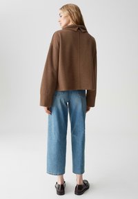 Maglione corto marrone con colletto ripiegato, abbinato a jeans a gamba larga di un leggero blu. Il modello indossa scarpe nere, mostrando una superficie liscia.