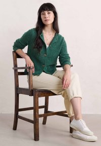 Zelený vlněný kardigan s texturovaným povrchem, s knoflíkovým zapínáním a přiléhavými rukávy, doplněný o béžové kalhoty a bílé tenisky.