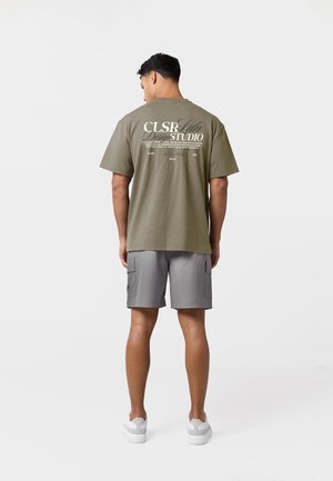 Bărbat purtând tricou cu grafică verde măsliniu și pantaloni scurți cargo gri, stând cu spatele spre cameră pe fundal alb.