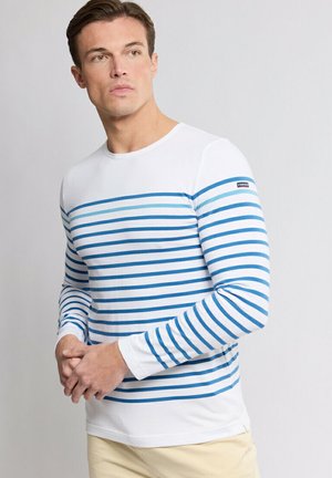 Homme portant une chemise blanche à manches longues avec des rayures horizontales bleues, debout les mains jointes, regardant légèrement vers la gauche.