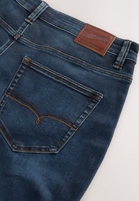 Dark blue denim jeans with orange stitching, visible back pocket, belt loops, and a brown leather patch labeled "Premium Denim Goods."