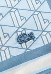 Lyseblåt stoff har et geometrisk mønster med hvite aksenter. Et lite, brodert design av en krokodille i mørkeblått er synlig.
