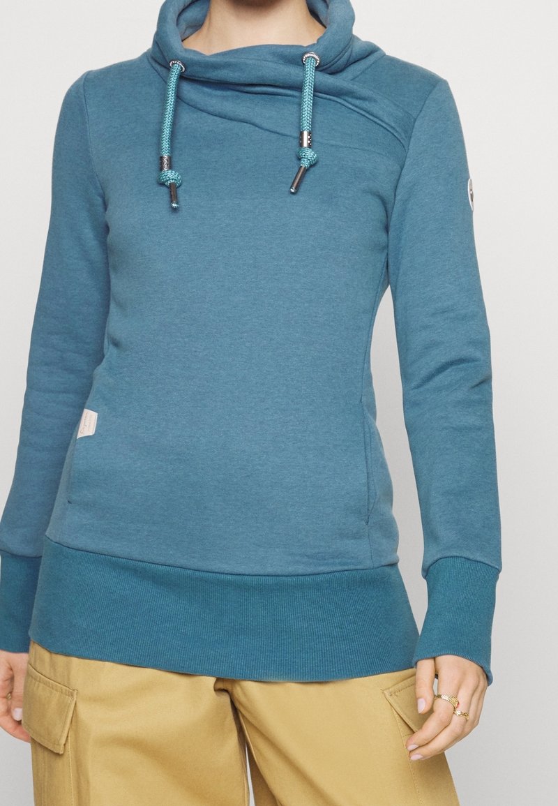 Personne portant un sweat à capuche turquoise à manches longues avec cordons et un pantalon cargo beige, debout devant un fond clair uni.