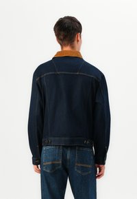 Donkerdenim jack met een bruin corduroy-kraag, zichtbare stikdetails en knoopaccenten op de taille en manchetten. Model gezien vanuit de achterkant.