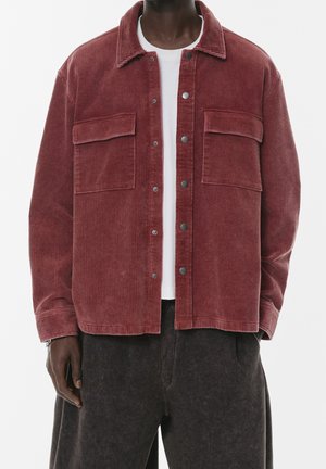 Homme portant une veste en velours côtelé couleur rouille avec des boutons pression, par-dessus un t-shirt blanc et un pantalon foncé, debout devant un fond blanc.