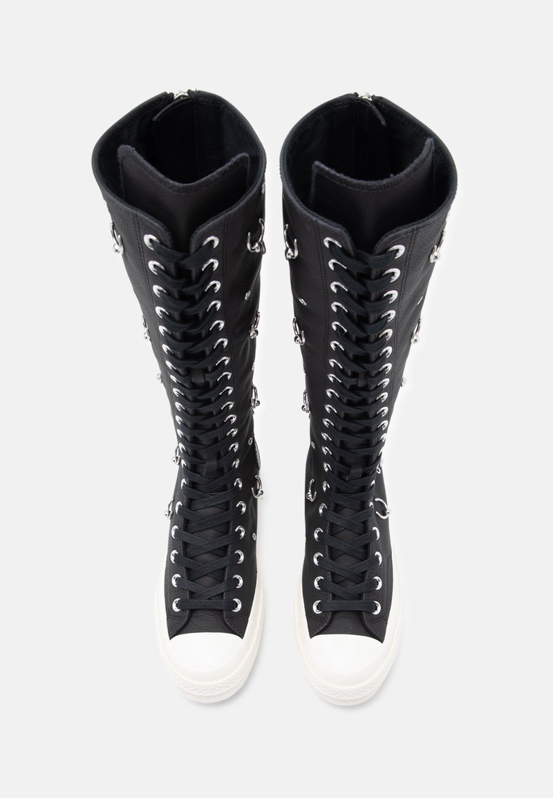 Svarta höghöga stövlar med snörning fram, silverfärgade ögla och en vit gummisula. Överdelen har en slät textur och en strukturerad design.