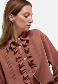 Blouse marron à volants avec un devant boutonné, présentant des détails de couture verticaux et un col structuré. Texture de tissu lisse.