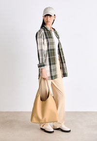 Femme portant une casquette blanche, un haut blanc, une chemise à carreaux verte, un pantalon beige, des chaussures crème, tenant un grand sac à bandoulière marron clair sur un fond uni.