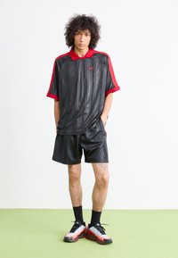 Young person with curly hair wearing black and red Nike sportswear, standing with hands in pockets on light green floor against white wall.