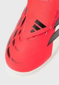 Κοντινό πλάνο σε ένα κόκκινο αθλητικό παπούτσι με μαύρα τριγωνικά σχέδια και μαύρο λογότυπο Adidas στο λουράκι.