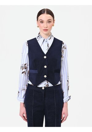 Donna con capelli pettinati all'indietro che indossa orecchini a cerchio dorati, una camicia a fiori a righe, un gilet blu navy con bottoni e pantaloni coordinati.
