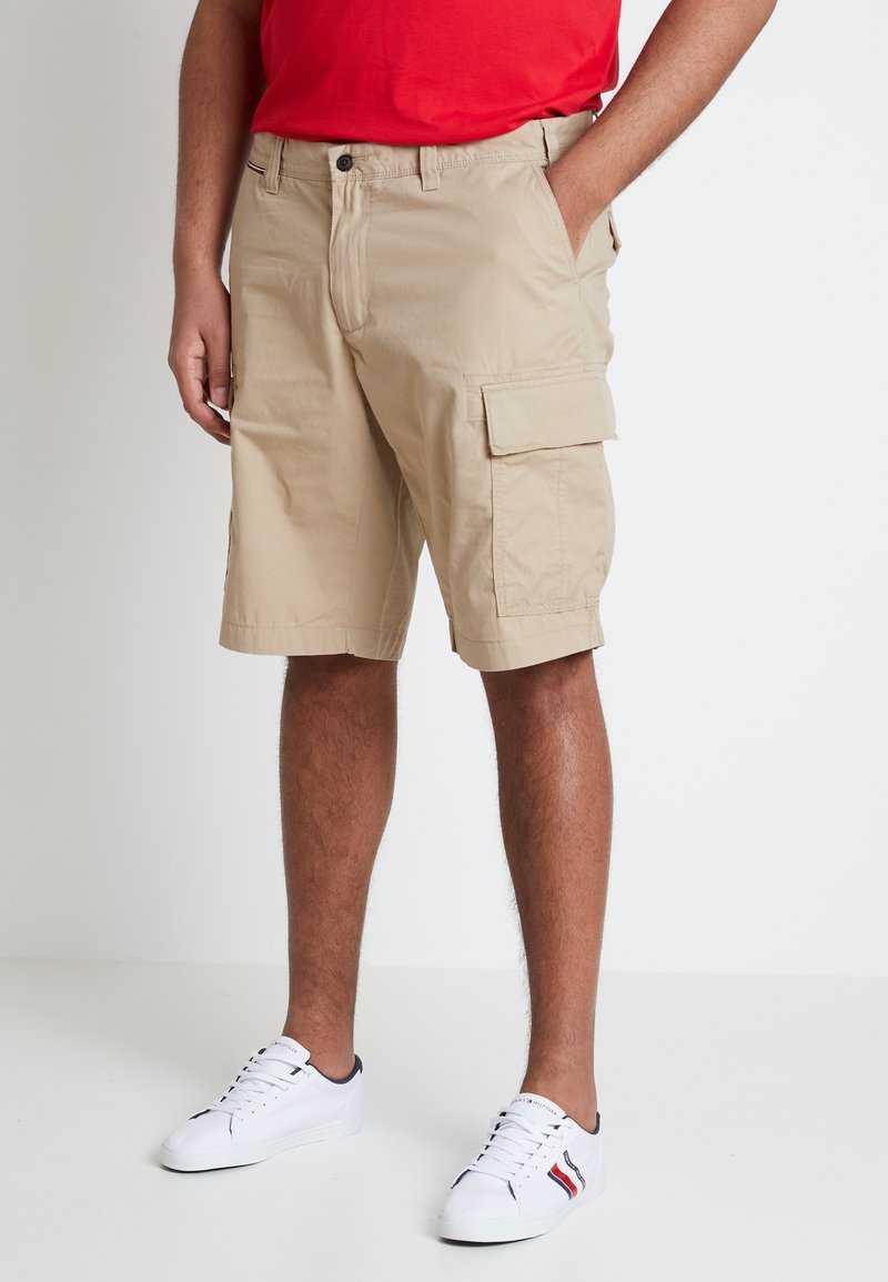 Personne portant un short cargo beige, des baskets blanches avec des détails rouges et bleu marine, et un t-shirt rouge, debout devant un fond uni.