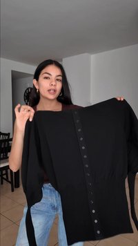 Žena s dlouhými tmavými vlasy drží černý zapínací cardigan s nařaseným pasem, stojící v minimalistickém vnitřním jídelním prostoru.