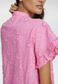 Blusa rosa ricamata con motivi floreali, maniche a scallop e tessuto testurizzato. Il design presenta dettagli di cucitura intricati.