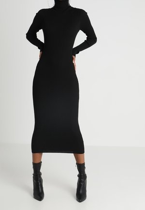 Frau trägt ein eng anliegendes, langärmeliges schwarzes Midi-Kleid mit Rollkragen und schwarzen Stiefeletten, steht mit den Händen in der Hüfte vor einem weißen Hintergrund.