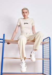 Beige cropped t-shirt with black "Dickies" logo, paired with cream corduroy pants. White sneakers with black accents. Sitting on a blue metal shelf.