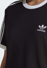 Černé bavlněné tričko s bílým kulatým výstřihem, které má tři bílé boční pruhy a malé bílé logo Adidas na levé hrudi.