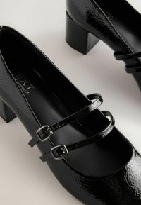 Black patent leather Mary Jane shoes with a square toe, featuring two adjustable straps and a mid-height block heel. Textured surface detail.