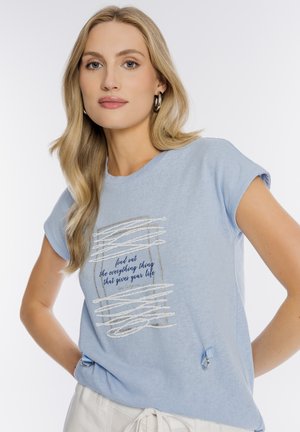 Blondynka w jasnoniebieskiej koszulce z napisem „dowiedz się wszystkiego, co nadaje sens twojemu życiu,” w połączeniu z białymi spodniami.