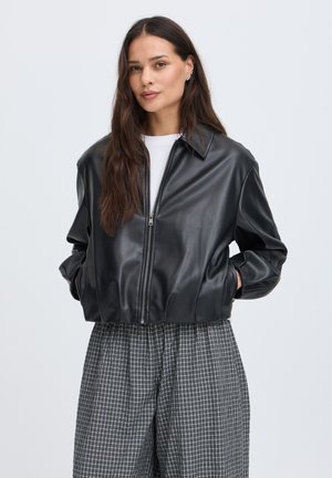 Femme aux longs cheveux bruns portant une veste en cuir noire zippée, un top blanc et un pantalon large gris à carreaux, les mains dans les poches.