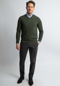 Groene v-hals trui, lange mouwen, gedragen over een lichtblauw shirt. Donkergrijze broek en donkere schoenen. Eenvoudig, casual ontwerp zonder patronen.