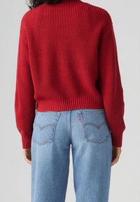 Henkilö, joka on pukeutunut punaisiin ribattuihin neuleisiin ja vaaleansinisiin korkeavyötäröisiin farkkuihin, näkyy selkäpuolelta yksinkertaista taustaa vasten.