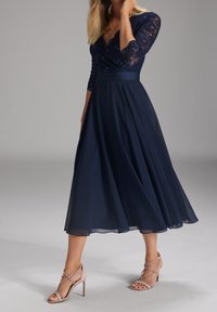 Marineblaues Kleid mit einem Spitzenoberteil und drei Viertel-Ärmeln. Fließender A-Linien-Rock, kombiniert mit hellrosa Riemchenheels.