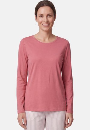 Frau mit braunen Haaren zu einem niedrigen Dutt gebunden trägt ein langärmeliges, staubrosa Shirt und hellrosa-weiß gestreifte Hosen und lächelt in die Kamera.