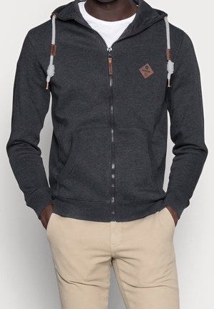 Man wearing dark gray zip-up hoodie with white drawstrings and beige pants, hands in pockets, against plain light background.