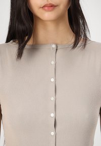 Haut beige à côtes avec un col rond et une patte de boutonnage centrale. Le tissu est doux avec une texture subtile et possède six petits boutons blancs.