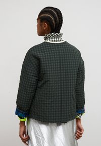 Person mit geflochtenem Haar trägt eine dunkelgrün karierte Jacke über einem silbernen Rock und steht mit dem Rücken zur Kamera vor einem einfarbigen Hintergrund.