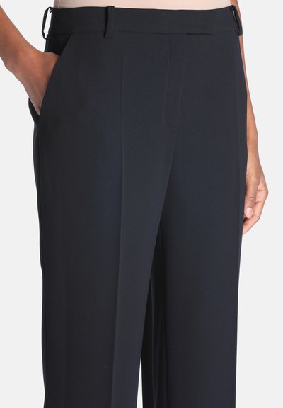 Gros plan d'une personne portant un pantalon noir sur mesure, la main dans la poche gauche et la main droite reposant sur le côté.