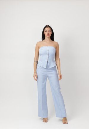 Vrouw staand, draagt een lichtblauwe strapless top met knopen en bijpassende wijde broekspijpen en doorzichtige hoge hakken tegen een effen achtergrond.