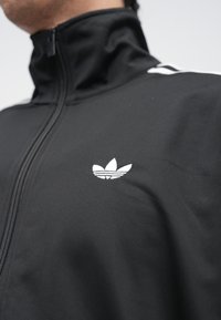 Čierna bunda na zips vyrobená z hladkej látky s bielym vyšívaným logom na hrudi a bielymi pruhmi na ramenách.