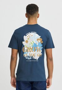 Marinblå bomullströja med blommönster, med texten "VÄX EFTERHAND" i ljusa och mörka nyanser. Korta ärmar, rund hals.